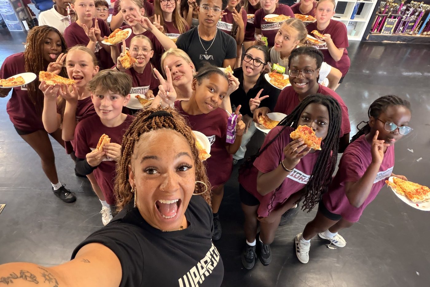 Warriorz dancers crowded together sharing pizza in the studio, trophies on the shelves behind