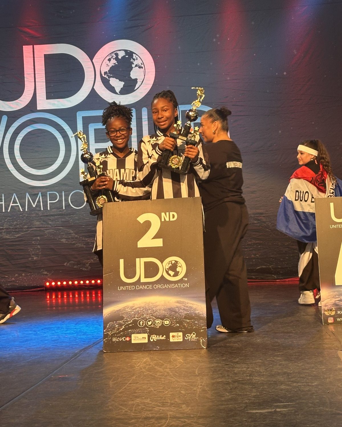 Two Warriorz dancers on stage at the UDO World Championships beside a second-place board, holding trophies
